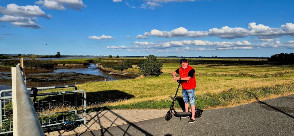 A Segway F2 Pro rider in a red jacket standing with his electric scooter on a path near the water.