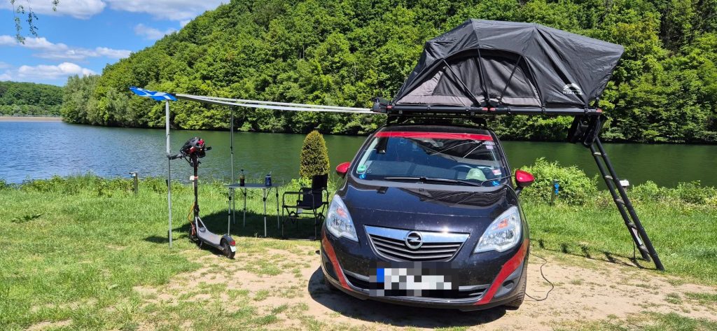 A car with a roof tent set up by a lake, with a Segway F2 Pro electric scooter parked beside the camp.