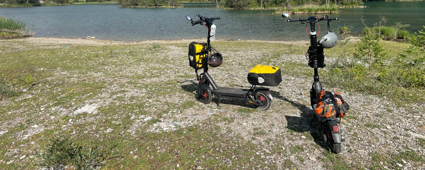 Two Segway MAX G3 electric scooters with travel bags and helmets parked by an alpine lake