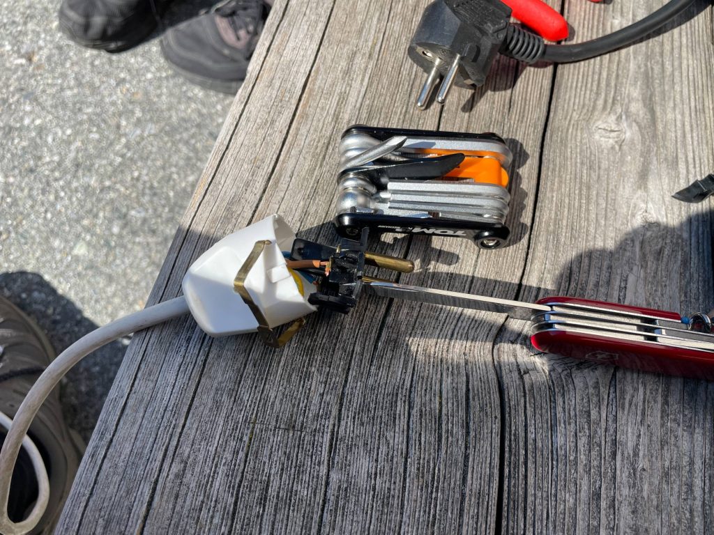 A charger plug being repaired with a multi-tool on a wooden bench during an alpine scooter trip
