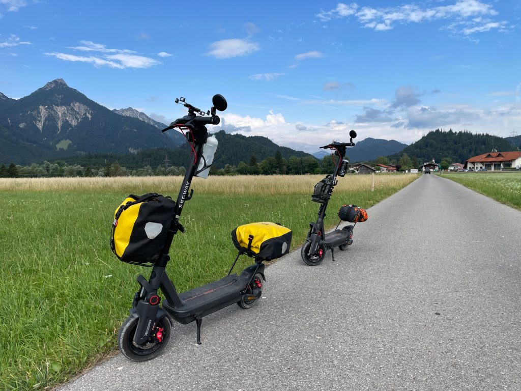 Two Segway MAX G3 electric scooters with rear-mounted waterproof bags on a country path below the Alps