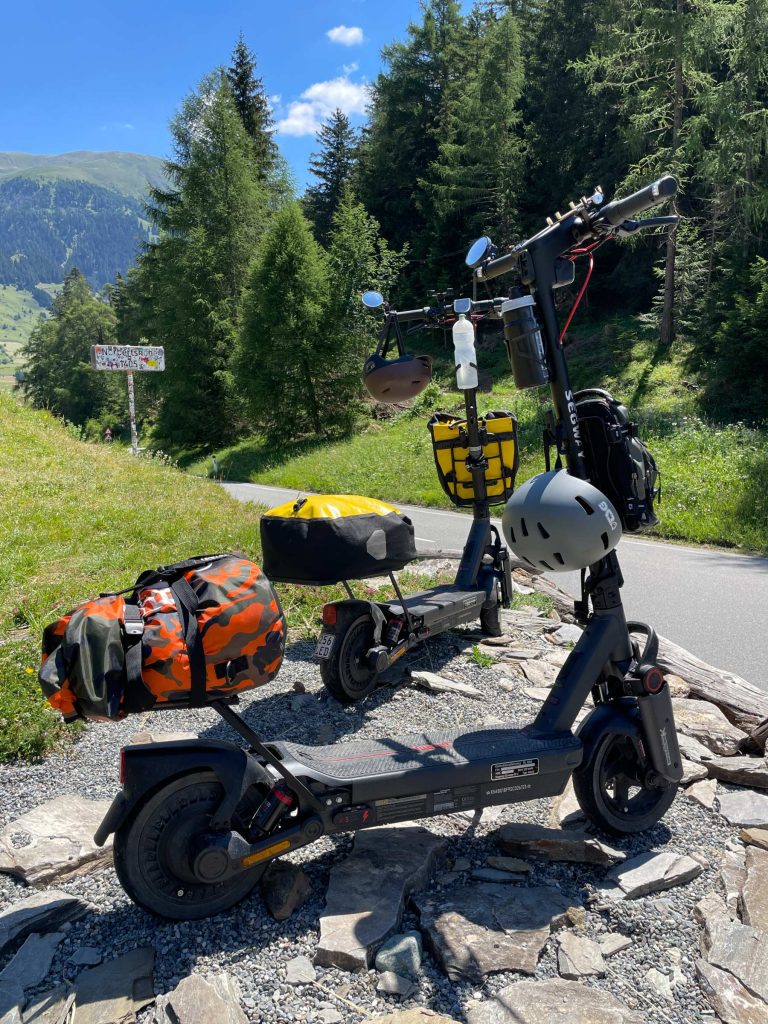 Segway MAX G3 electric scooter with travel bags and a helmet parked on gravel beside a mountain road