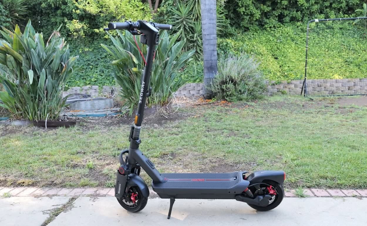 A Segway MAX G3 Plus electric scooter parked on a sidewalk next to a garden.