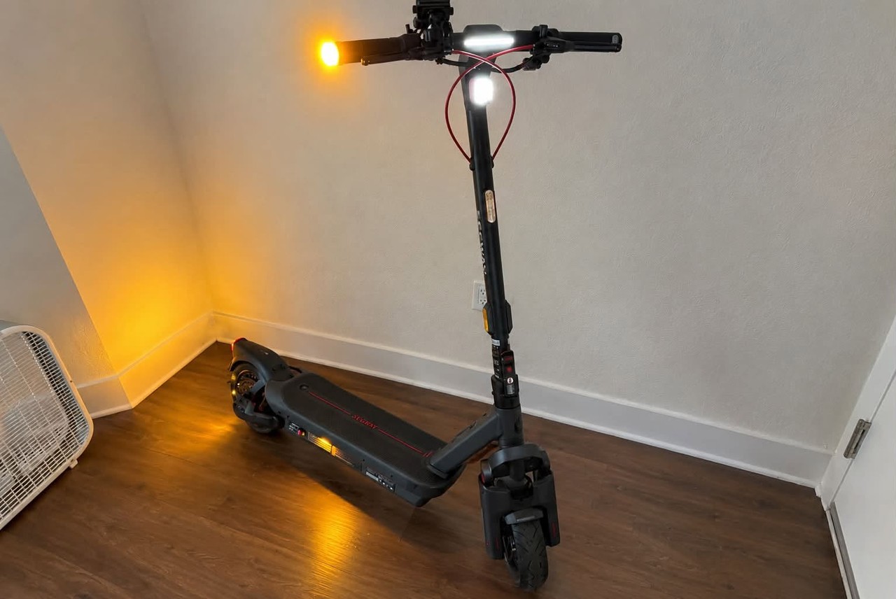 A Segway MAX G3 Plus electric scooter with its lights on, parked in the corner of a room.