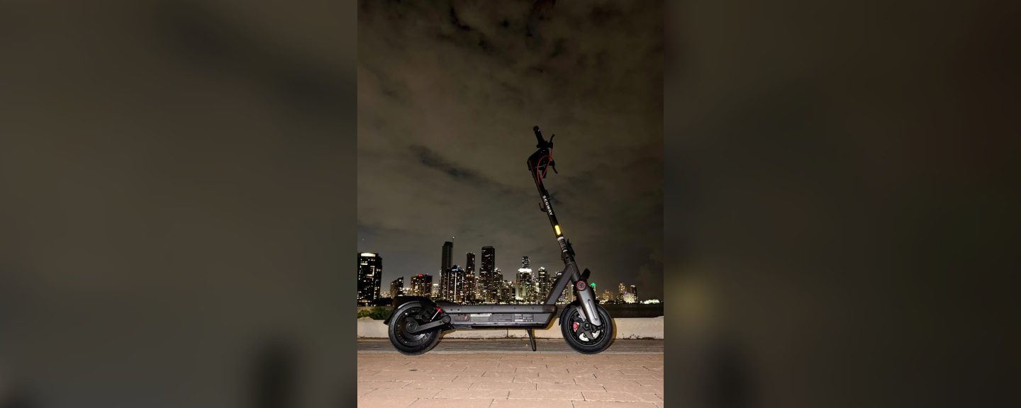 A Segway MAX G3 Plus electric scooter parked by the water with a city skyline lit up at night.