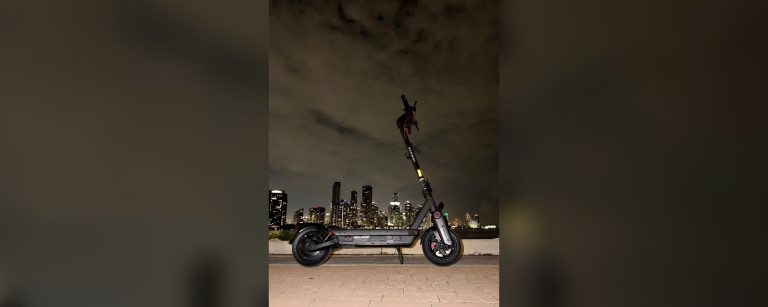 A Segway MAX G3 Plus electric scooter parked by the water with a city skyline lit up at night.