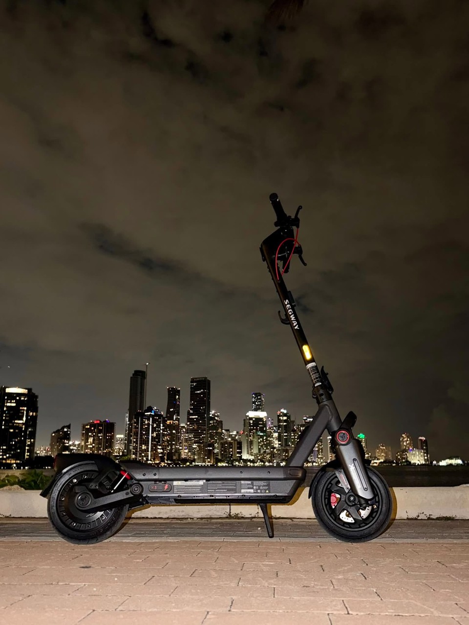 A Segway MAX G3 Plus electric scooter parked by the water with a city skyline lit up at night.