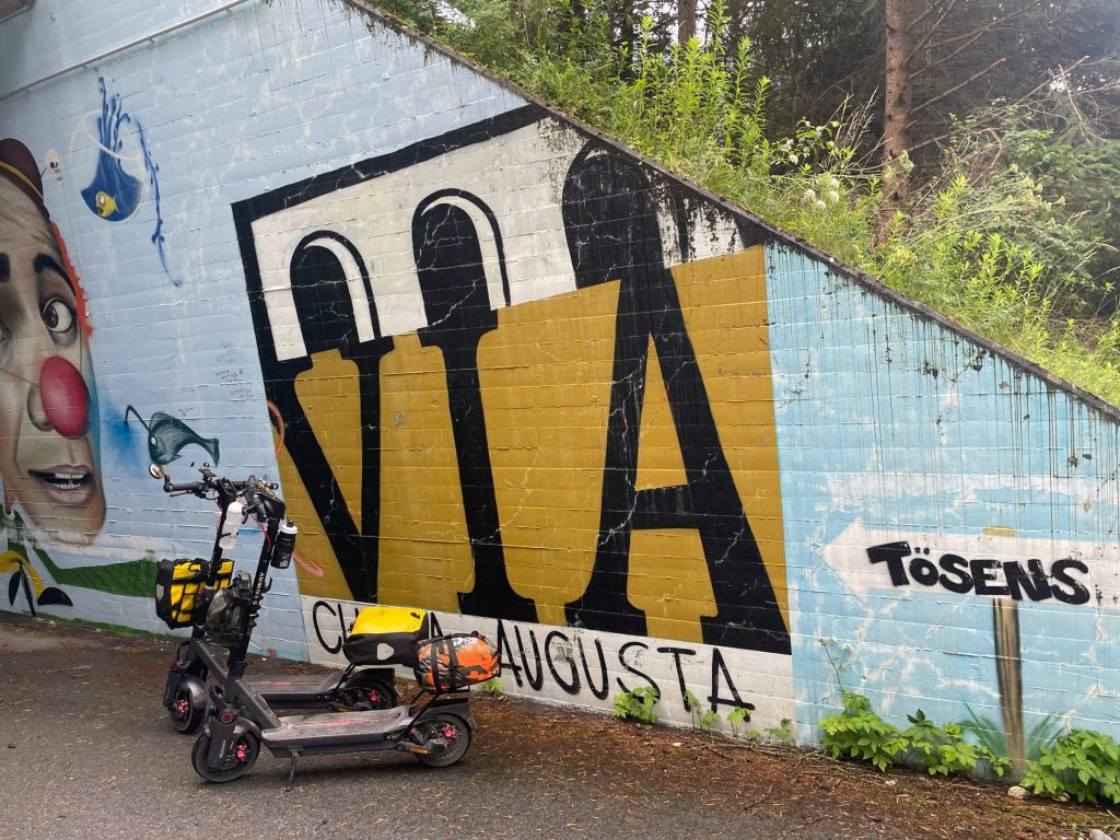Two Segway MAX G3 electric scooters loaded with bags parked beside a Via Claudia Augusta mural