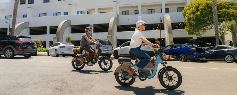 Two riders wearing helmets ride Segway MUXI Pro cargo eBikes down a sunny street in front of a white building